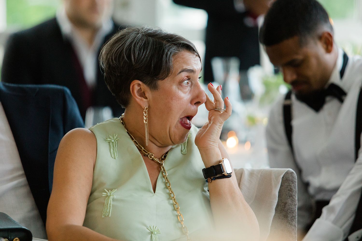 Wedding guest in a mint green dress reacts with surprise during reception while seated at table.