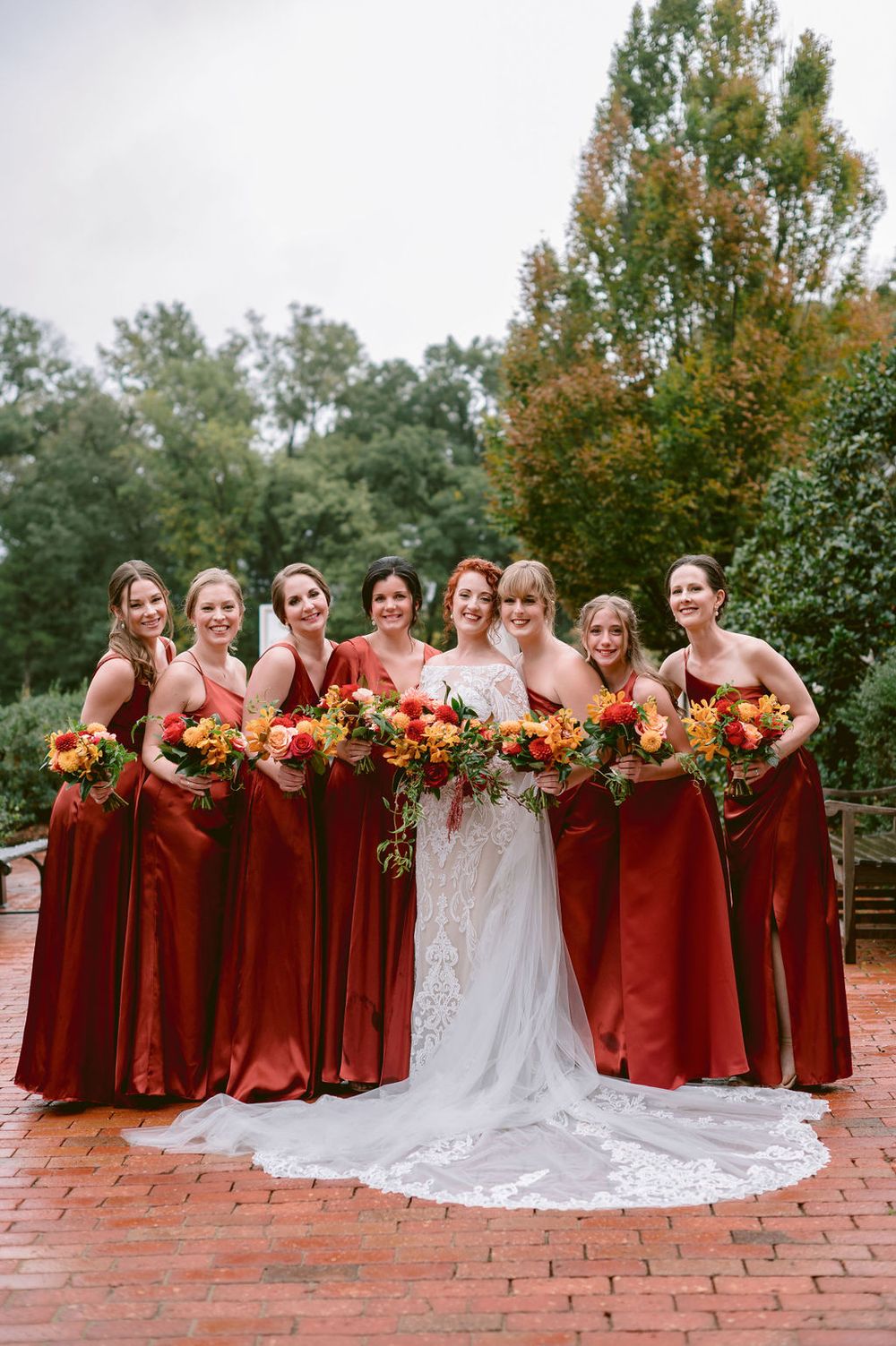 Wedding party in burgundy dresses and white gown pose together on brick patio with fall foliage backdrop.