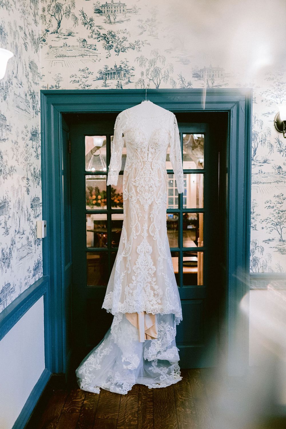 White lace wedding dress hanging in a room with blue-framed doorways and vintage toile wallpaper.