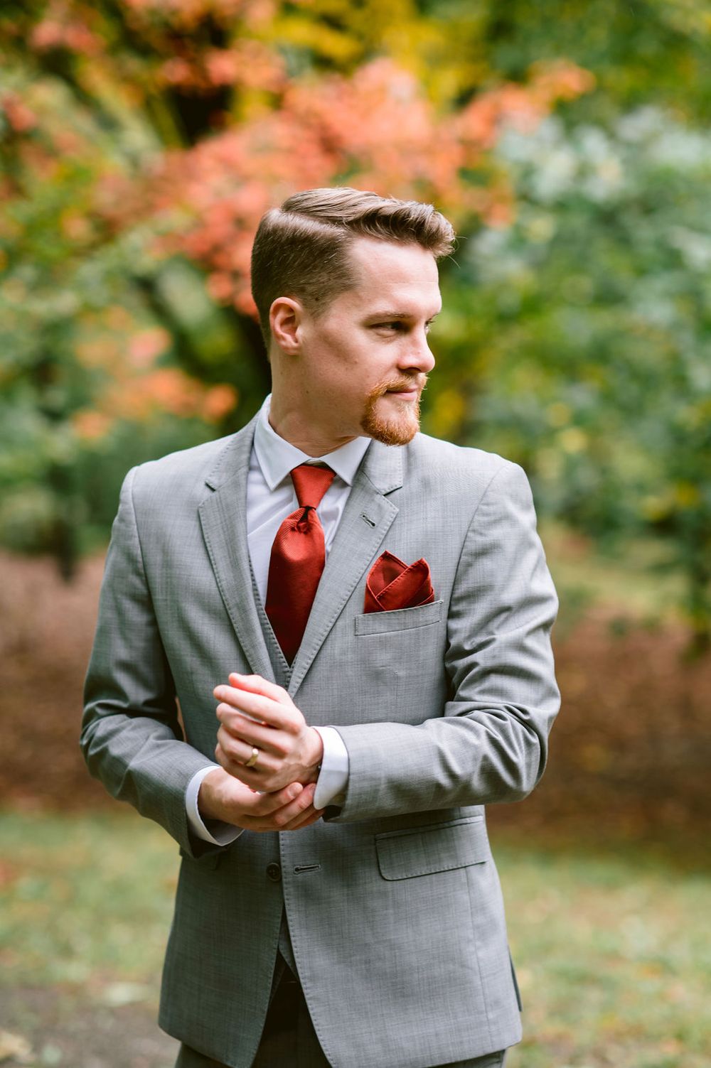 A person in a grey suit with red tie poses playfully in an outdoor autumn setting.