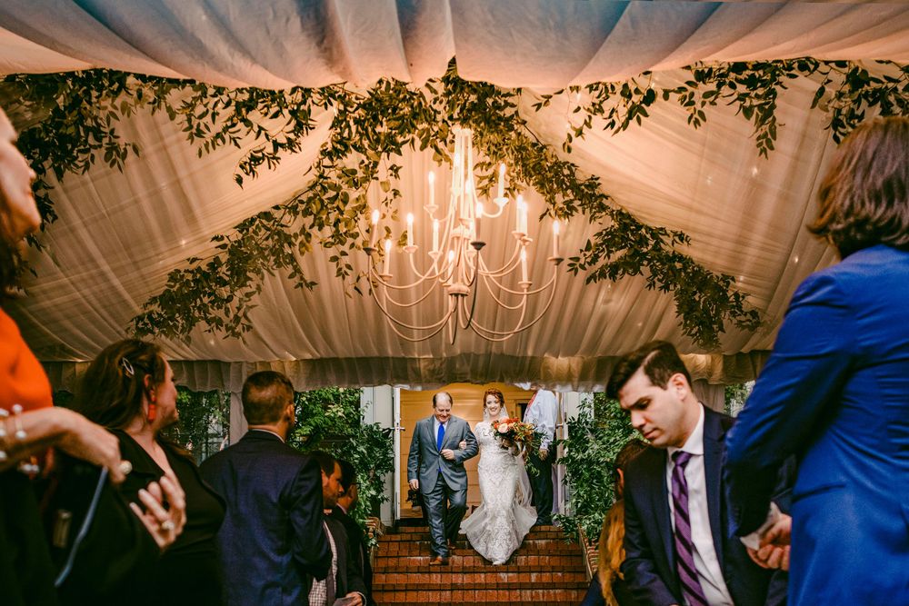 Wedding guests dance and celebrate on a dimly lit dance floor beneath elegant chandeliers and draped fabric decor.