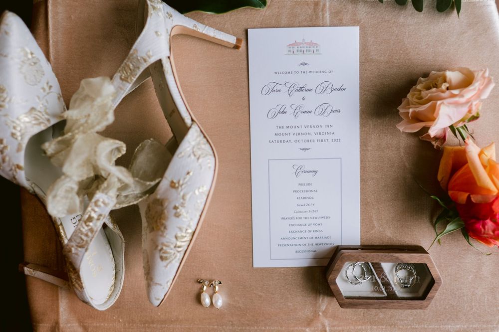 Elegant wedding details including invitations, rings, shoes, and floral elements arranged on a beige surface.