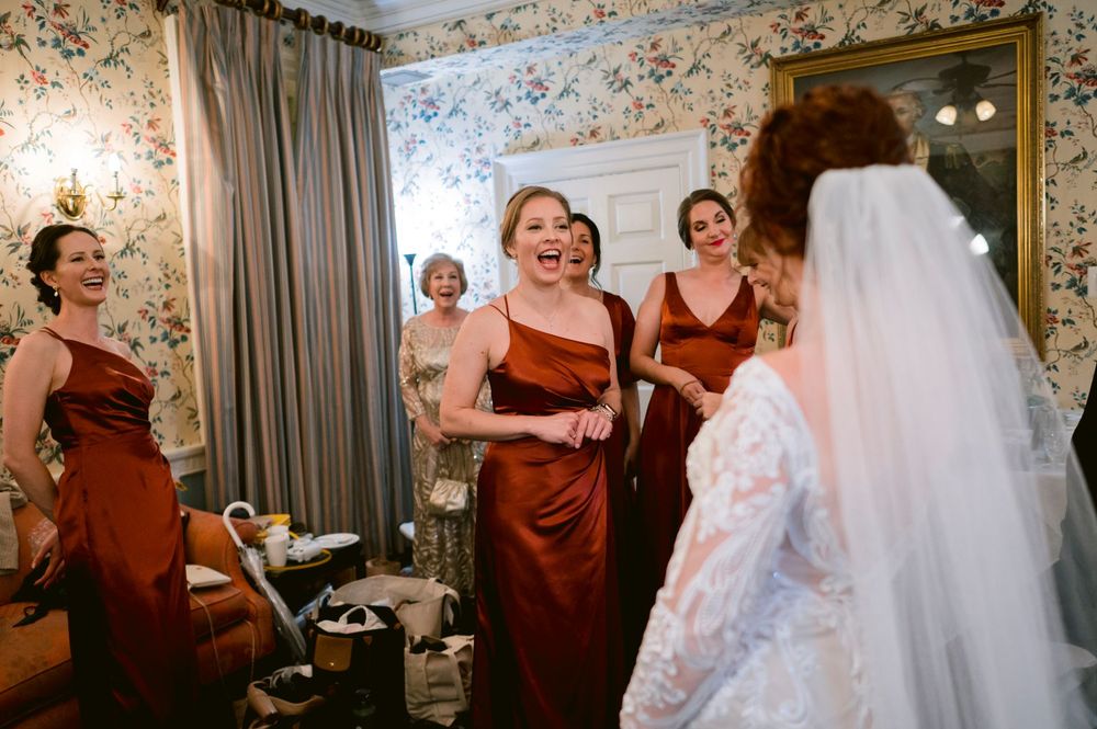 Wedding party members in burgundy dresses help bride in white gown get ready for the ceremony.