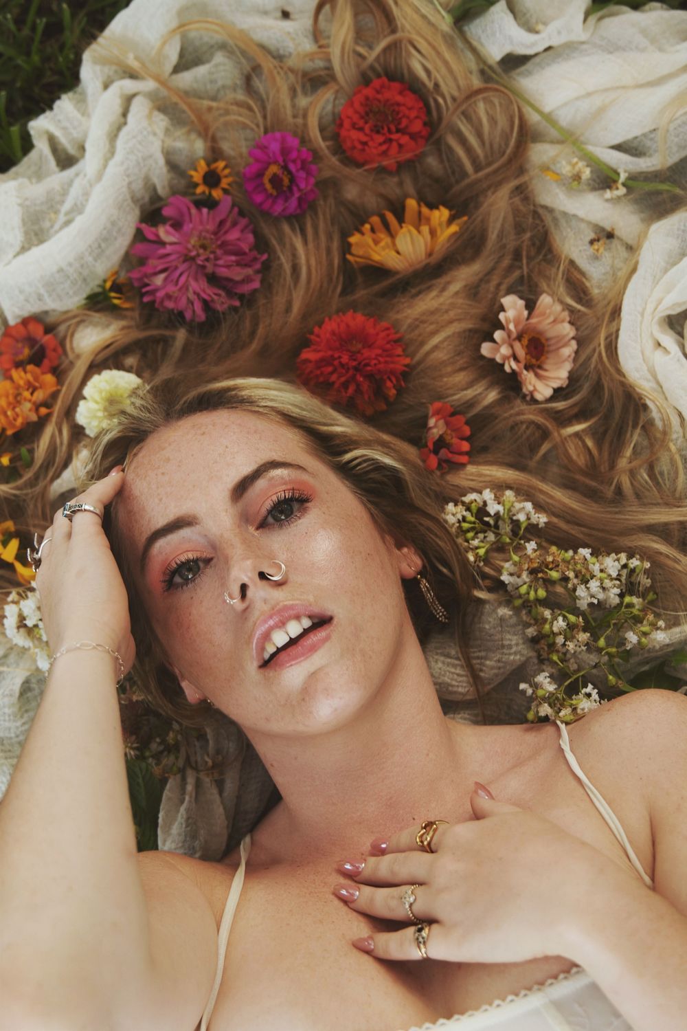 Artistic portrait of a person lying surrounded by colorful flowers and petals against a light background.