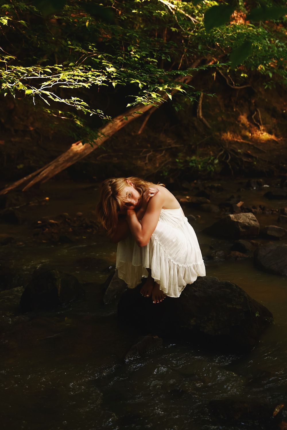 A figure in a white dress poses gracefully near dark water in a wooded setting with dramatic lighting and shadows.