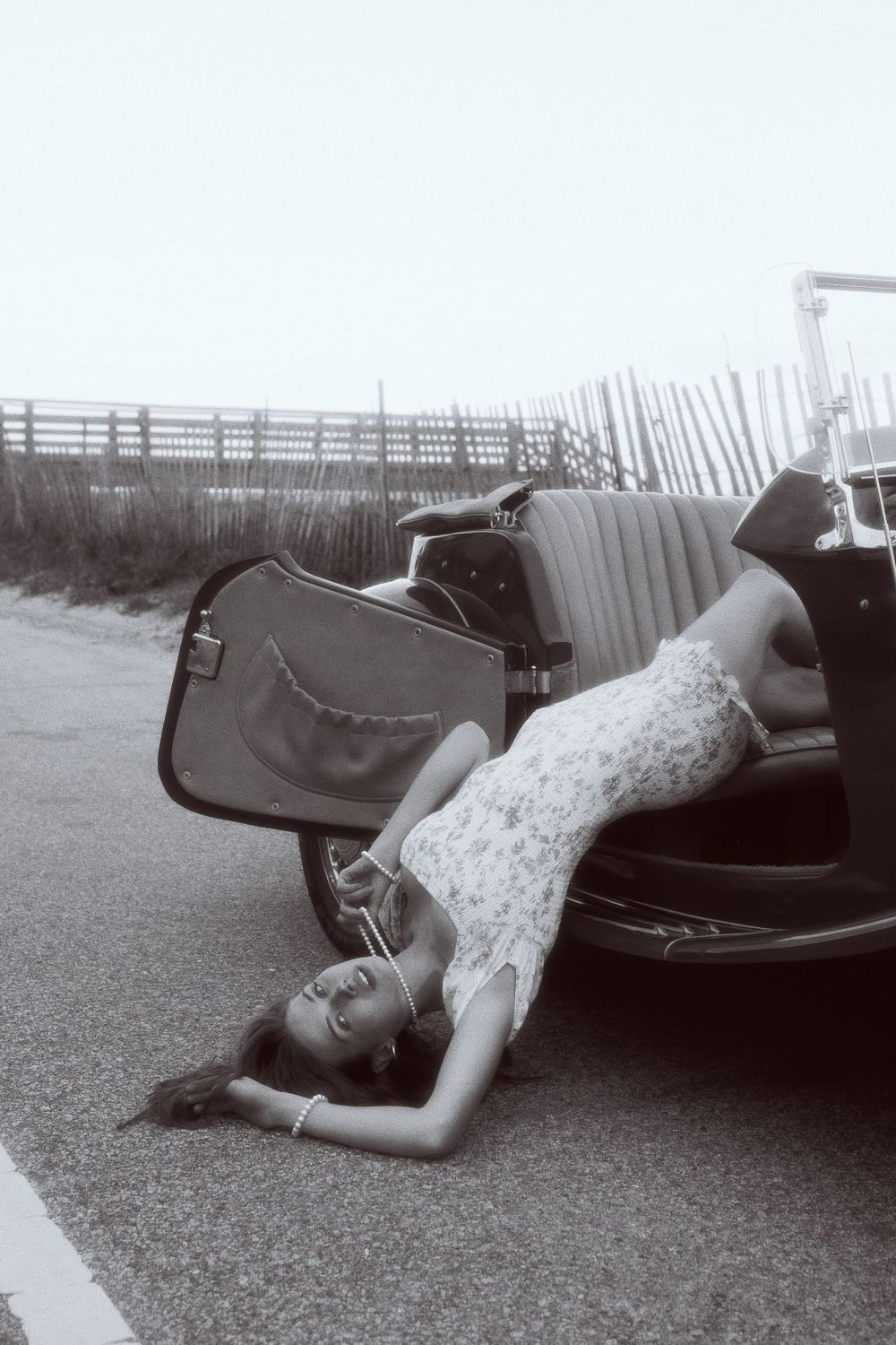 A person in a floral dress reclines dramatically across the back of a vintage car on a coastal boardwalk.