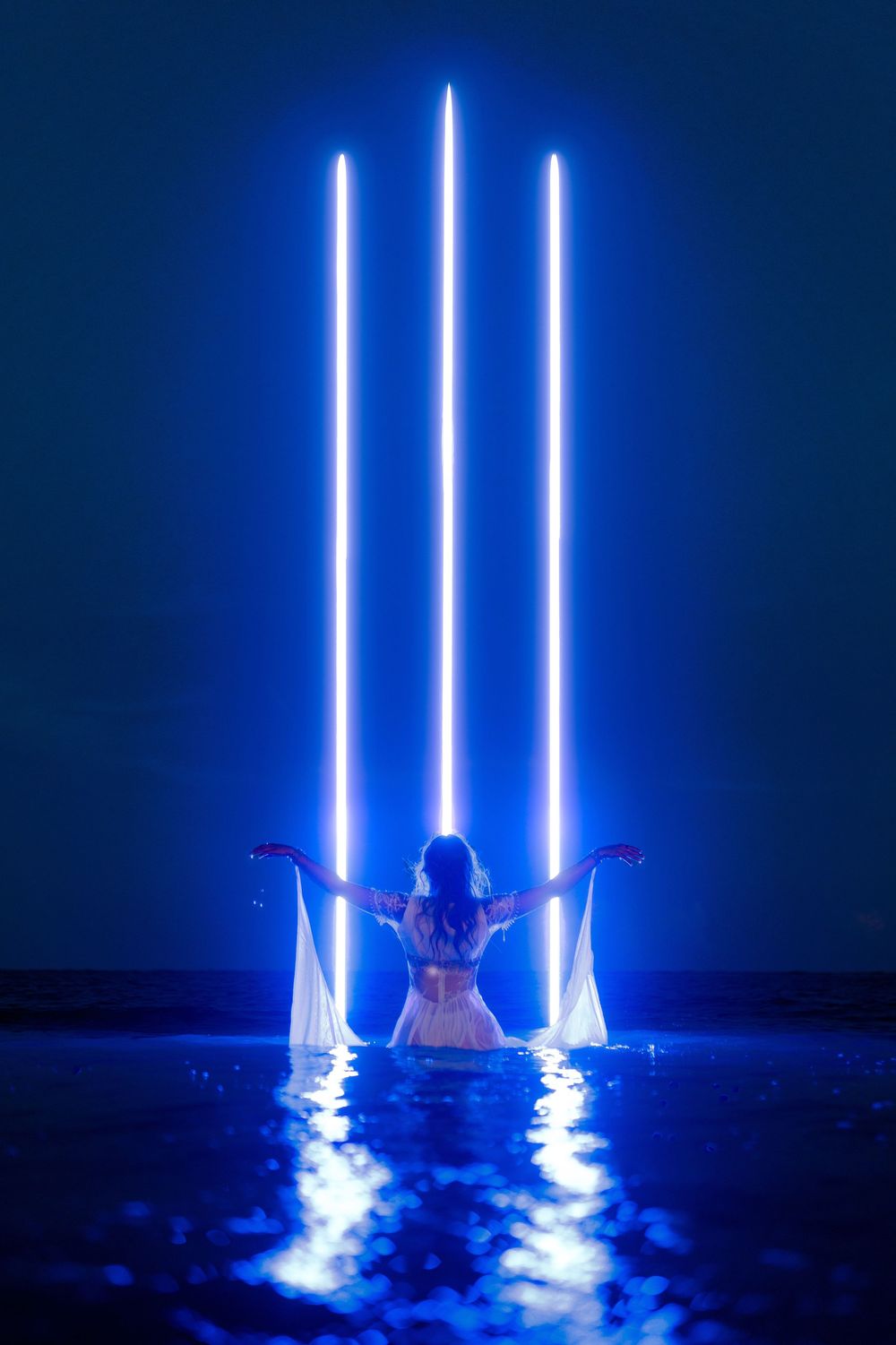 Silhouette figure in white dress stands in water beneath three glowing blue light beams creating ethereal reflections.