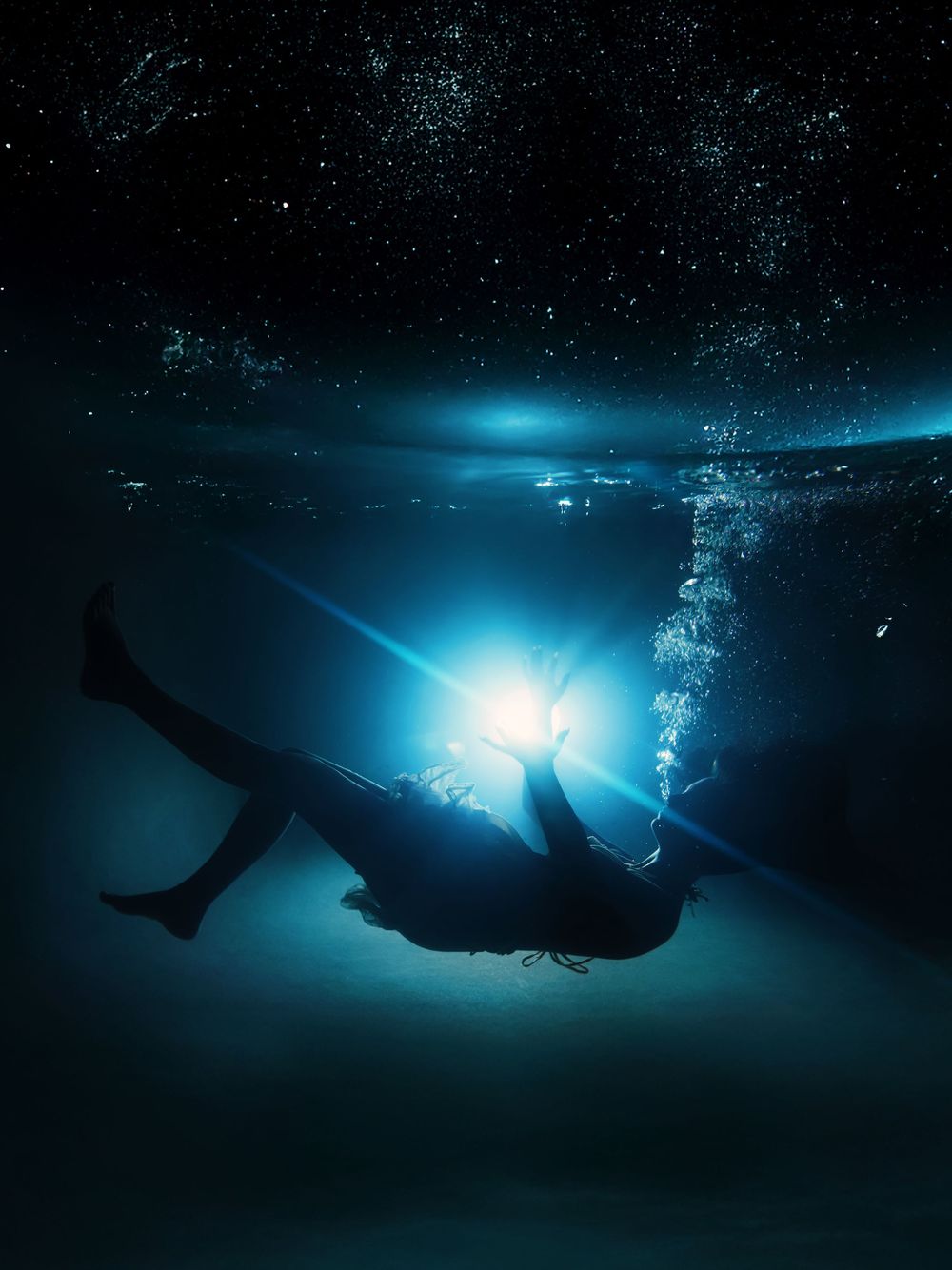 A silhouetted diver illuminated by bright underwater light creates a dramatic scene against deep blue ocean waters.