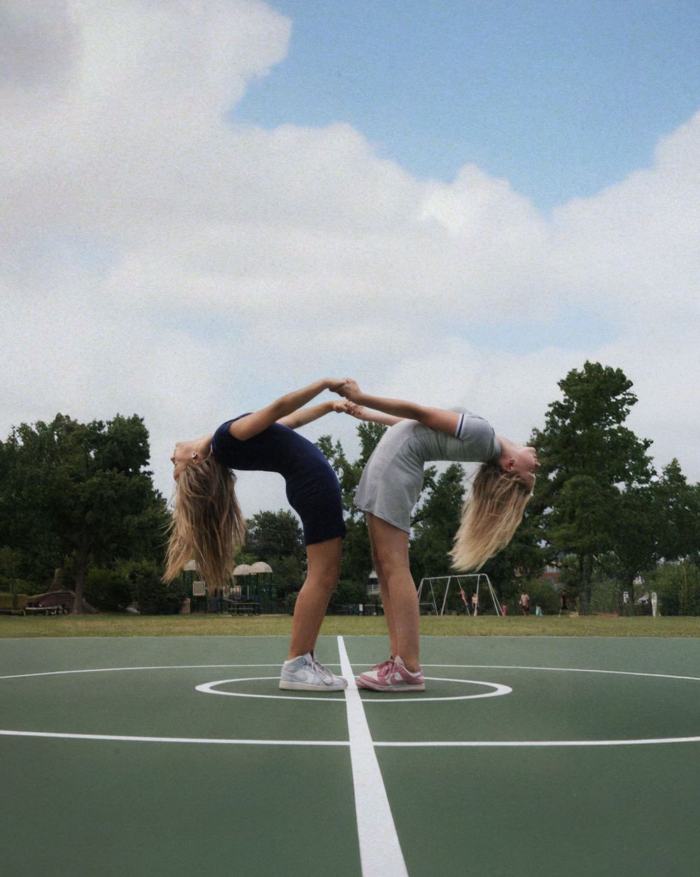Two people perform a synchronized backbend pose on a green basketball court under a blue sky with clouds.