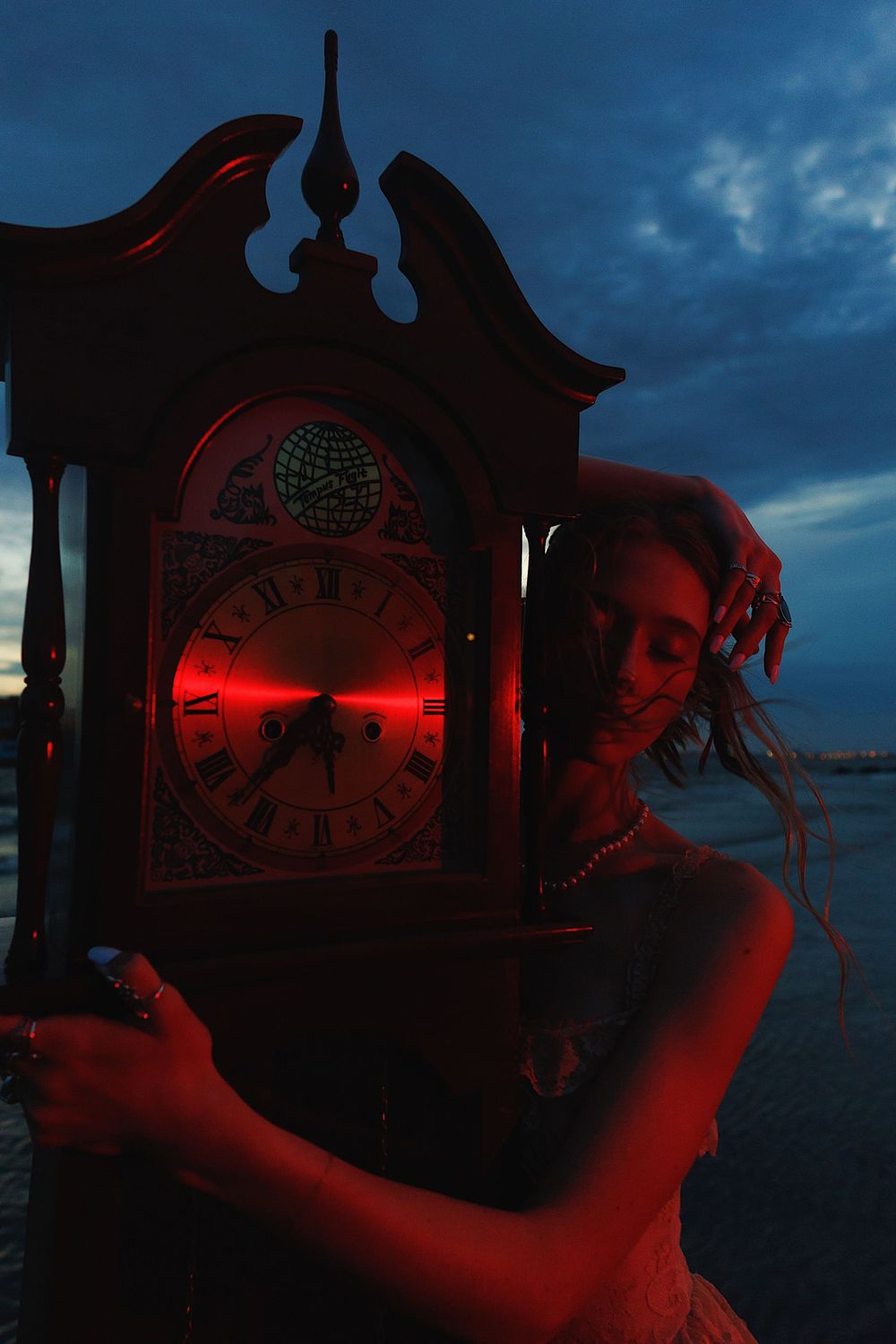 Vintage clock face illuminated in dramatic red lighting against dark blue evening sky.