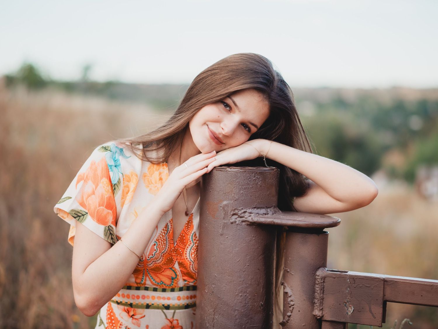 A rustic portrait series showing someone in an orange dress leaning against weathered wooden fence posts in nature.