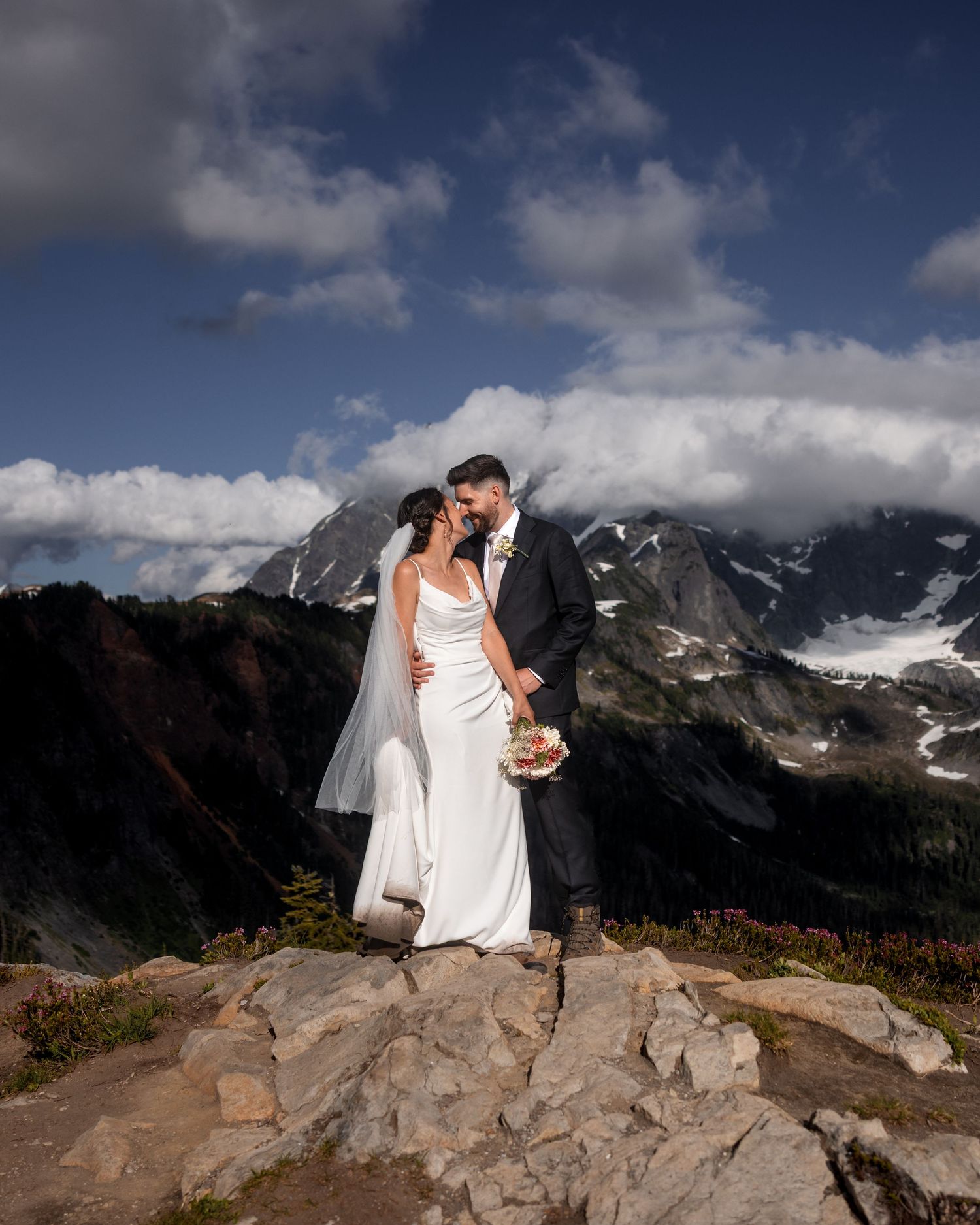 A wedding couple poses dramatically on rocky mountain peaks with snow-capped mountains in the background.