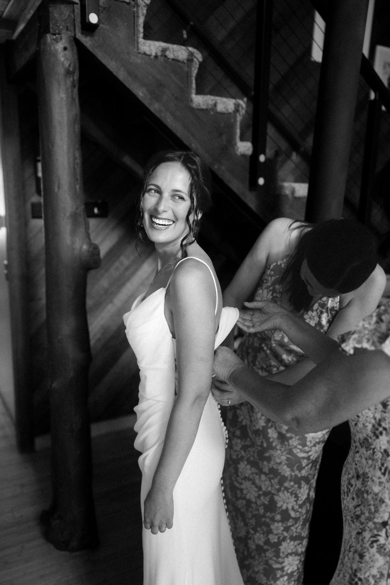 A candid black and white photo of a bride laughing while getting ready near rustic wooden stairs.