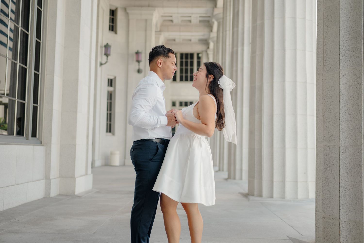 A couple shares a romantic moment in an elegant colonnade hallway with white pillars and natural lighting.