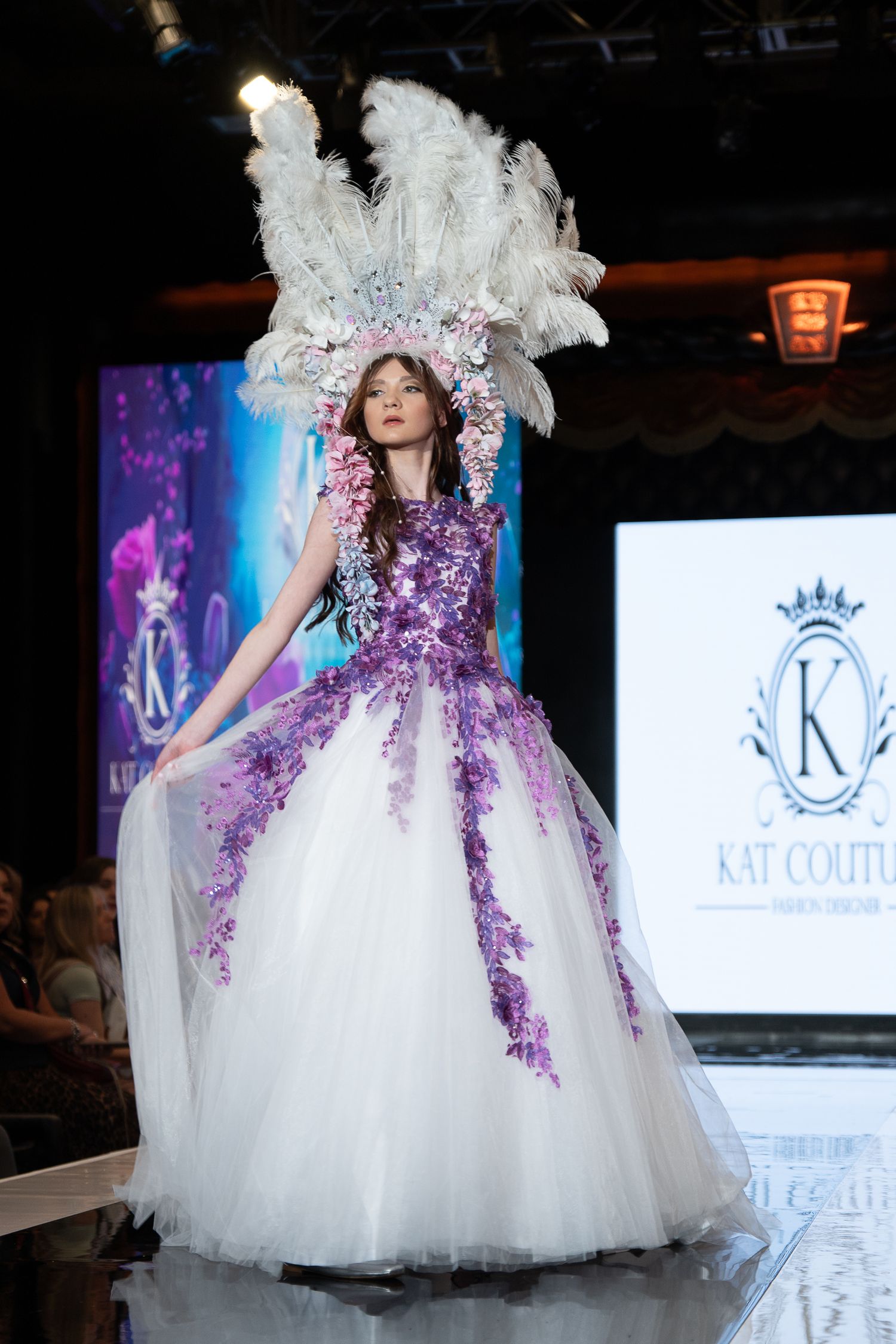 Elegant white and purple tulle ball gown with floral details showcased on a fashion runway with dramatic lighting.