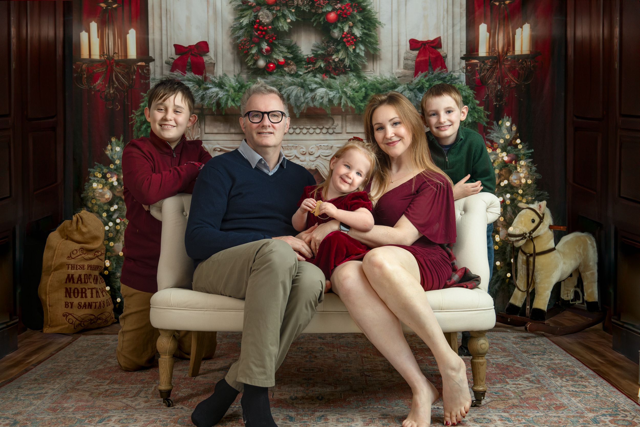 Family poses together on white couch in front of decorated Christmas tree and red wall during holiday photo session.