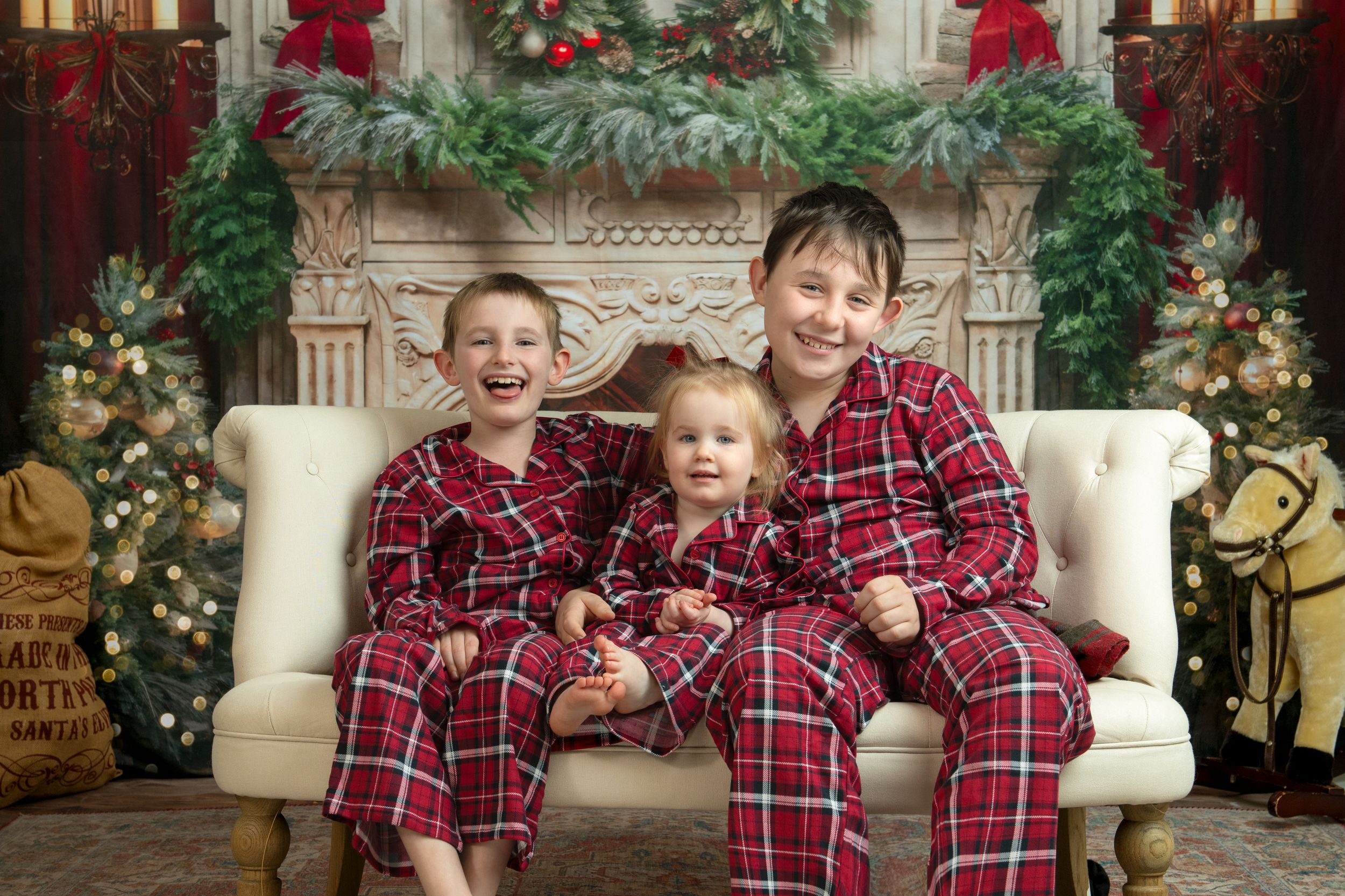 Three children in matching red plaid Christmas pajamas sit on a cream couch with holiday decorations in background.