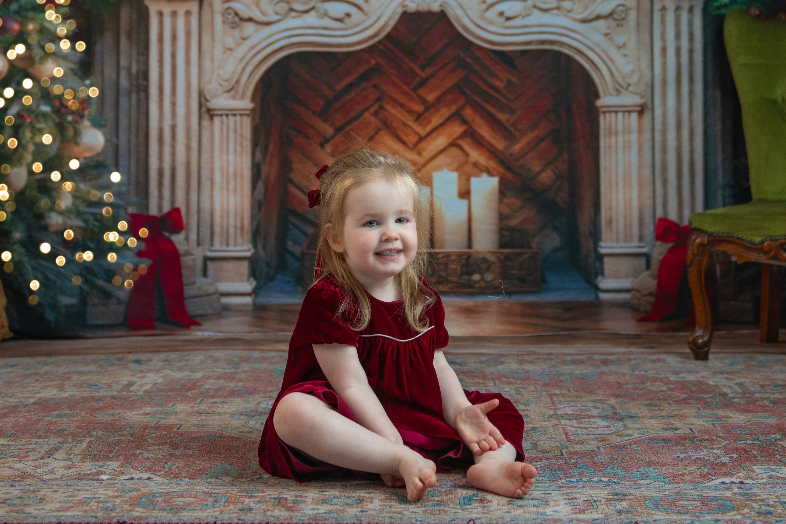 Child in a red velvet dress sits on ornate marble floor with Christmas tree and decorations in background.
