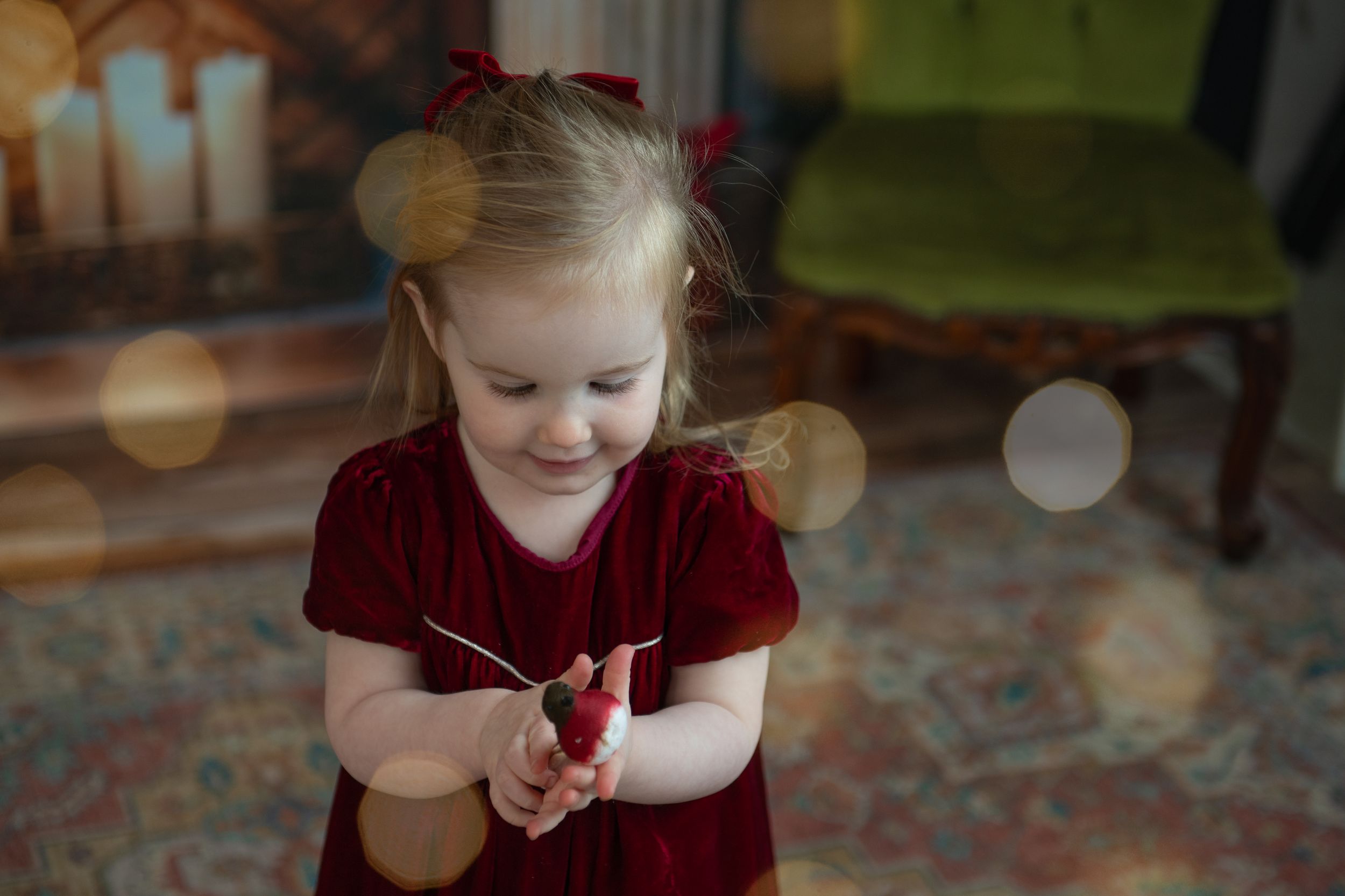 A child in a red velvet dress sits on a patterned carpet while playing with their hands.