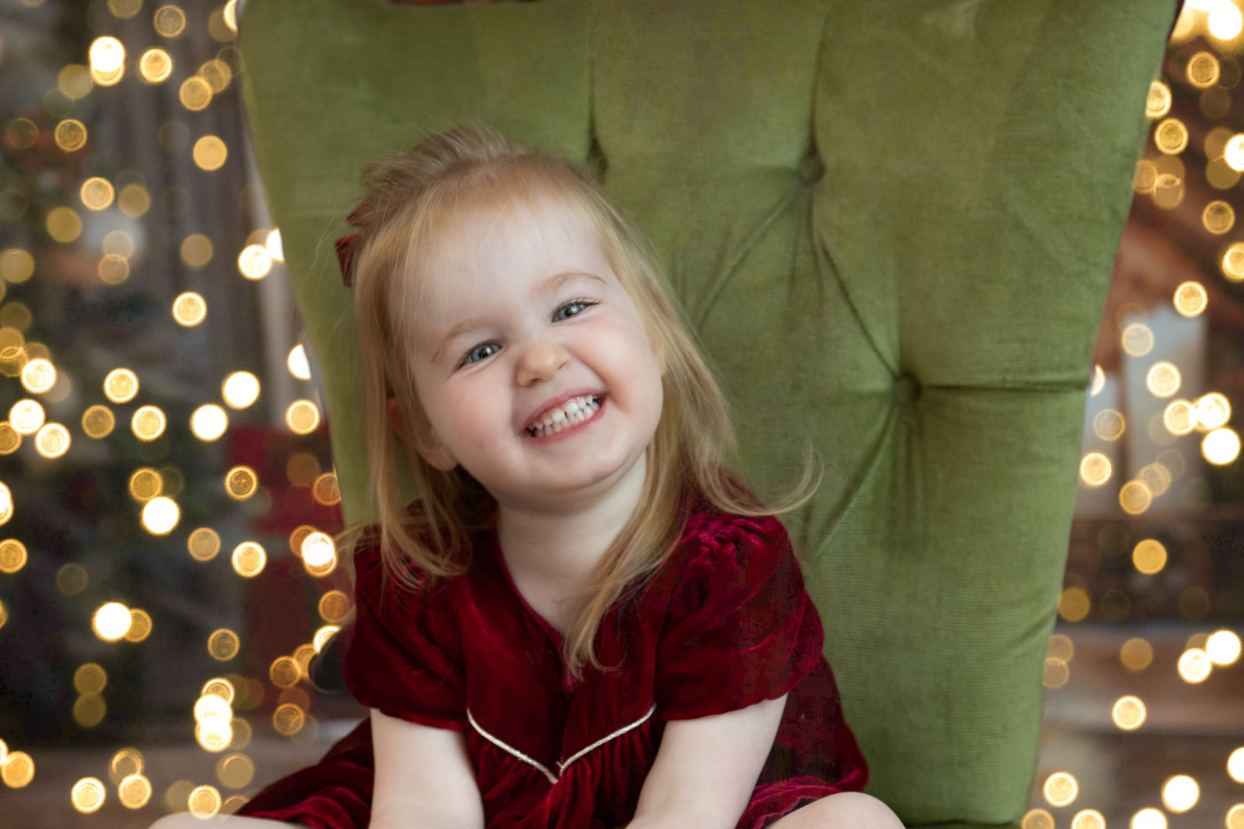 A child in red velvet dress sits on a green chair with twinkling holiday lights in the background.