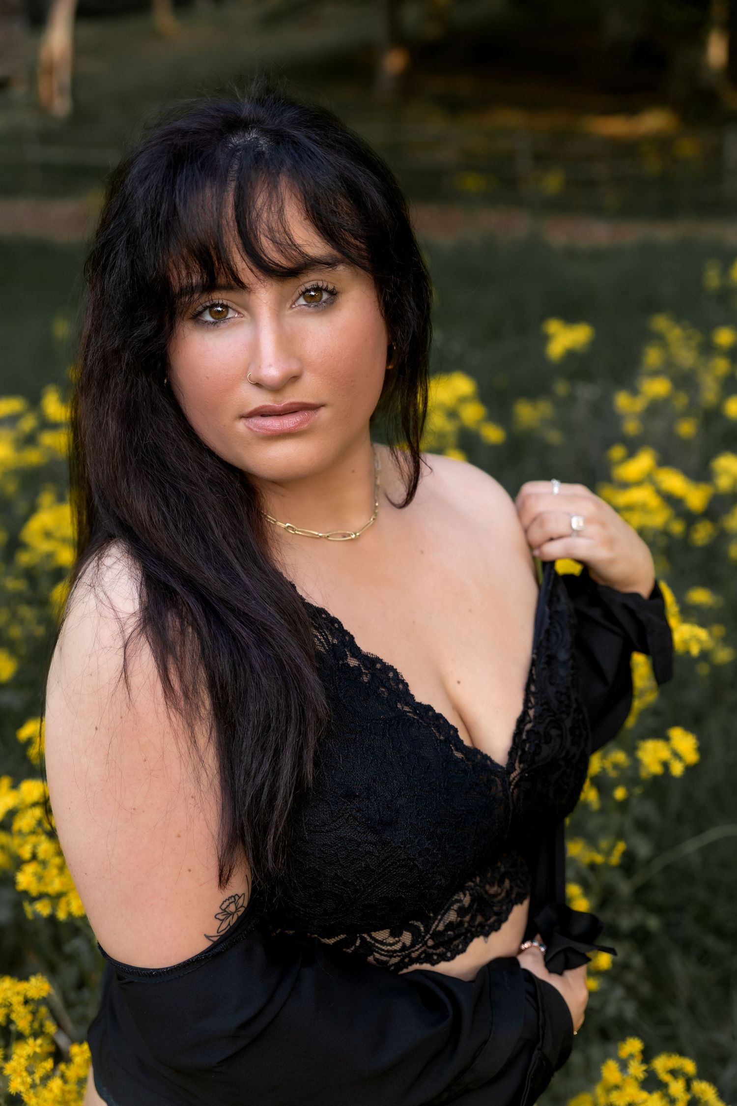 Woman with long dark hair poses in black dress among blooming yellow wildflowers in outdoor portrait series.