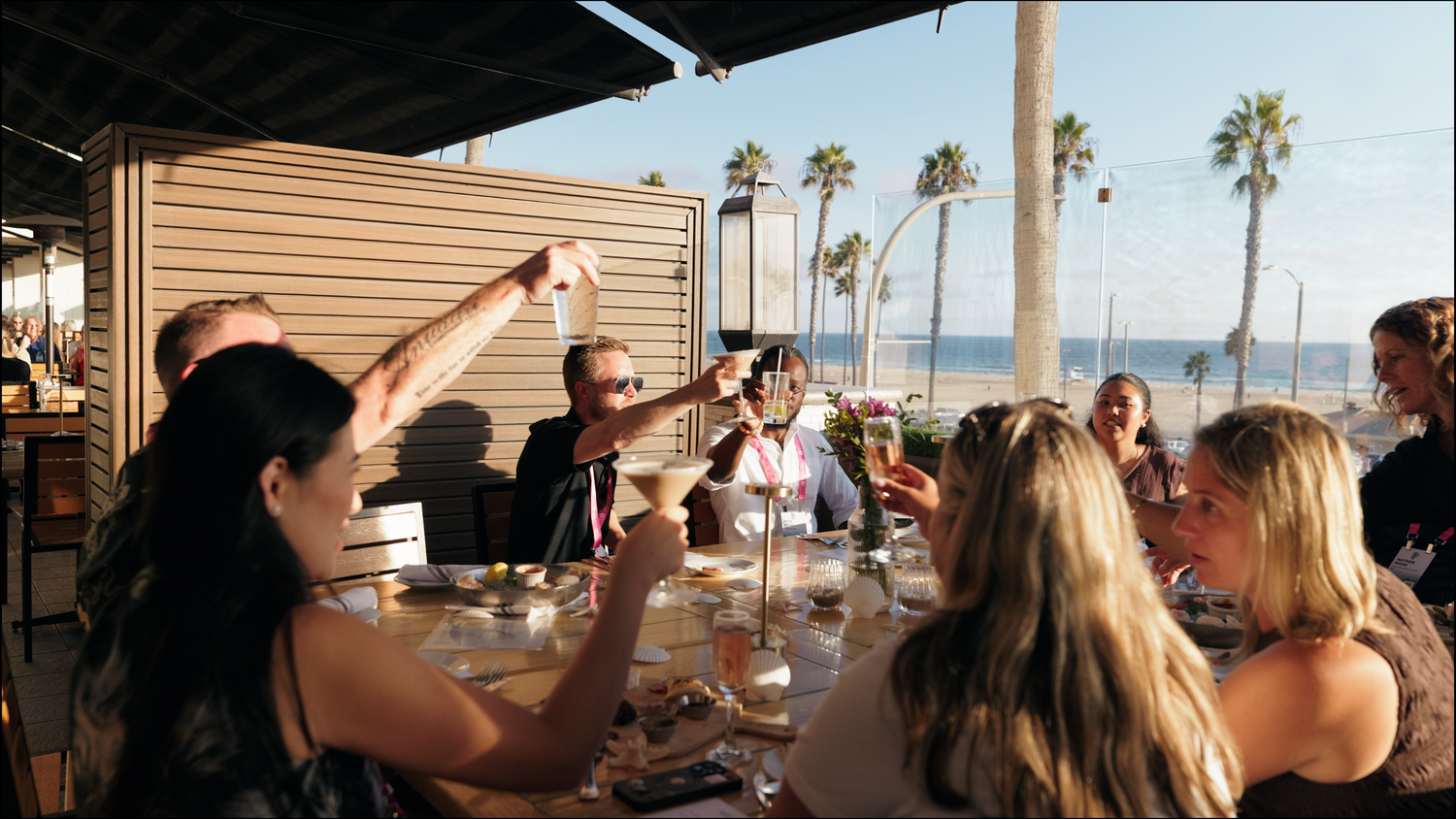 A group of friends raise their glasses in a toast at an oceanfront restaurant with palm trees in the background.