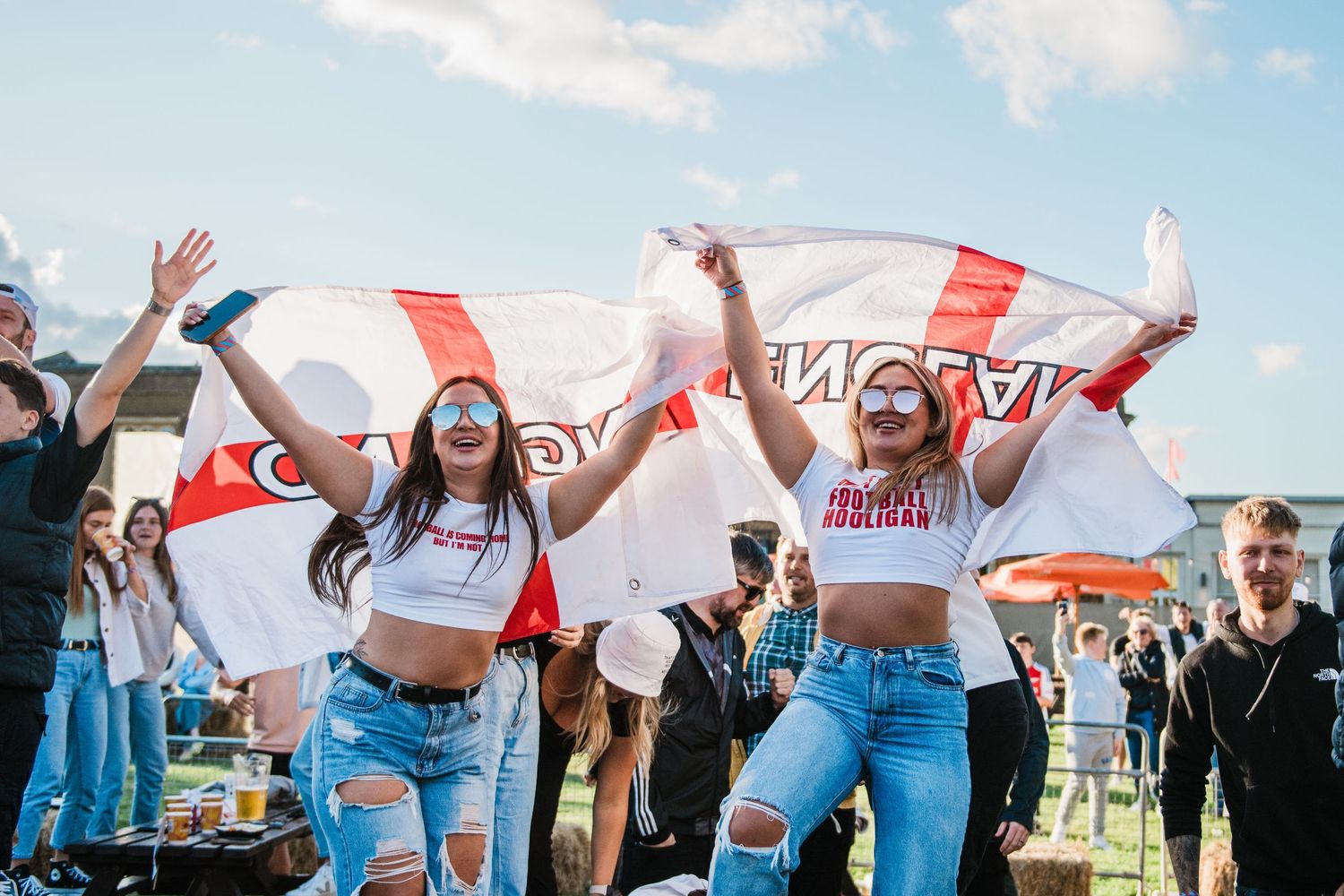 Two sports fans celebrate with England flags at an outdoor event wearing crop tops and ripped jeans.