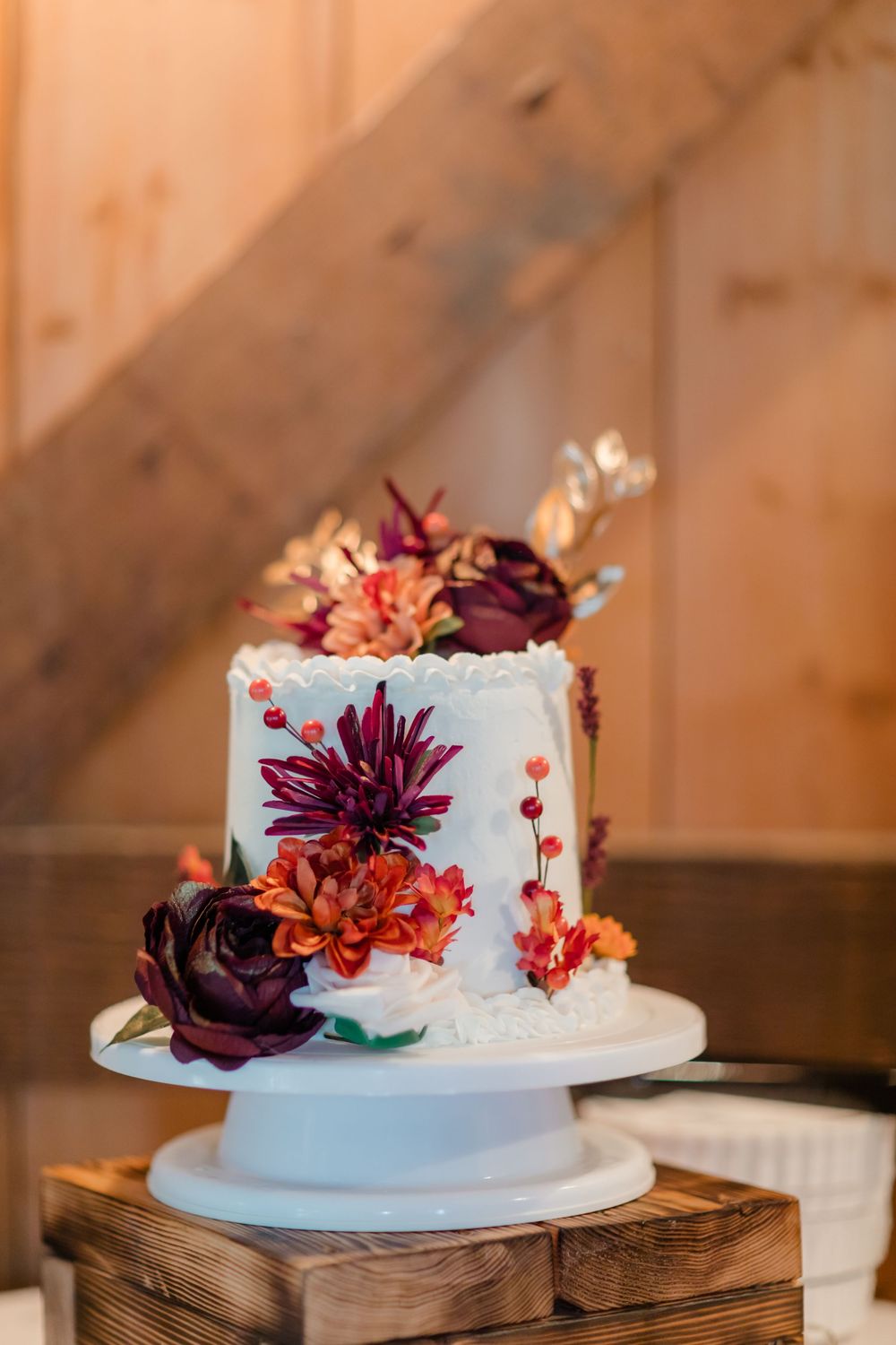 White wedding cake decorated with burgundy and orange flowers on a rustic wooden stand.