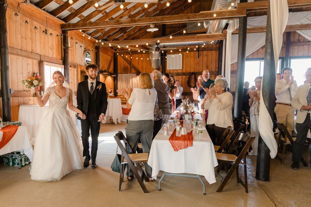 Wedding party celebrates in a rustic barn venue decorated with twinkling lights and elegant white decor.