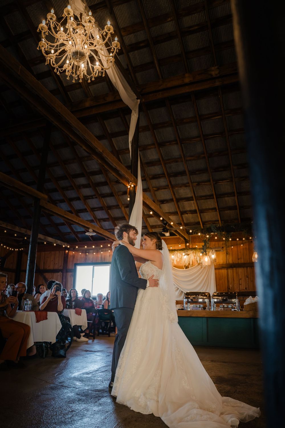 A romantic first dance between newlyweds under a rustic barn ceiling with twinkling lights and elegant chandeliers.
