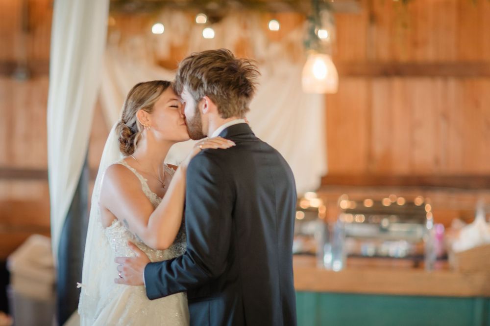 A couple shares their first dance in a rustic barn venue with warm lighting and wooden beams overhead.