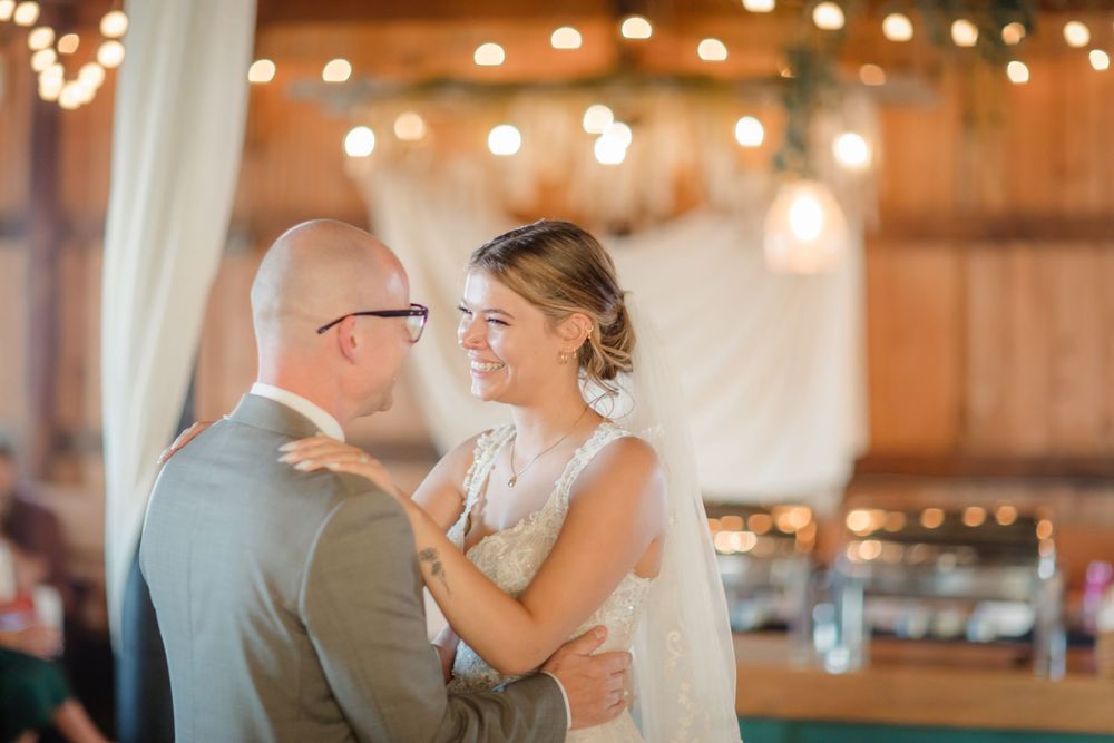A couple shares an intimate dance moment under warm string lights at a rustic wedding reception venue.