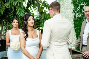 Wedding ceremony scene with bridesmaids in white dresses and tropical greenery backdrop.
