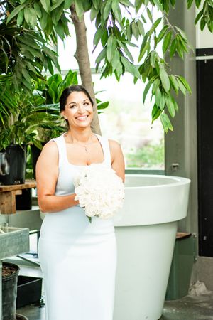 Bride in white dress poses with bouquet near modern bathtub in tropical setting.