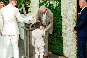 Wedding party members stand in white attire against a decorative floral wall backdrop.