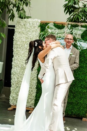 Wedding couple posing against a white floral wall and greenery backdrop.