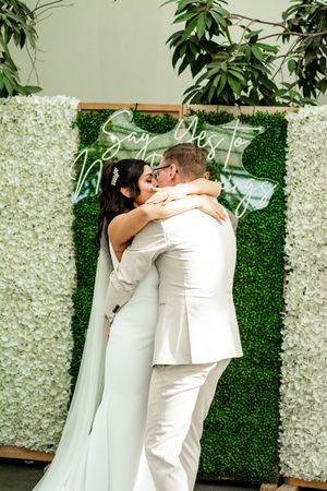 A romantic first kiss moment against a floral wall backdrop at a wedding ceremony.