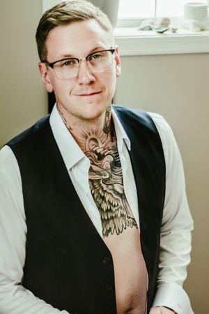 A person in glasses and vest with visible neck tattoos poses in natural window light.