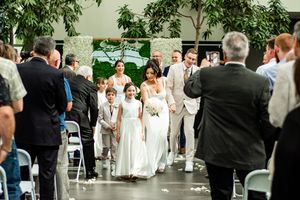 A wedding ceremony outdoors with guests seated in white chairs surrounded by greenery and trees.
