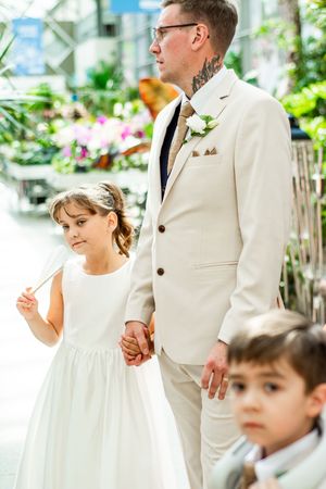 A groom in a cream suit walks with young attendants in a greenhouse setting filled with flowers and greenery.