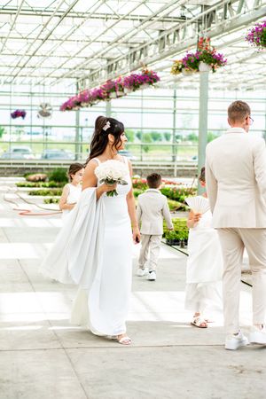 Wedding party walks through a greenhouse venue with hanging flower baskets and glass ceiling.