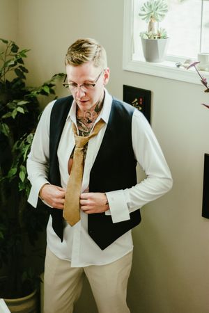 A person adjusts their gold tie while wearing a formal vest and white shirt in a sunlit room.