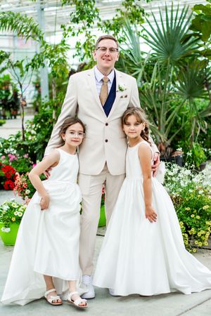 Children in white dresses and formal attire pose in a greenhouse garden setting.