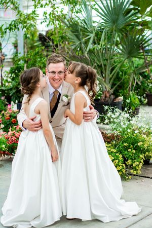 Two flower girls in white dresses share a sweet moment with a groomsman in a cream suit among lush garden plants.