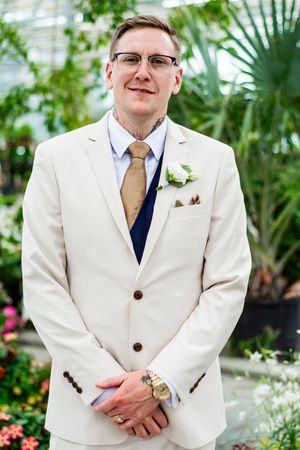 Groom in cream suit with navy tie and boutonniere poses in garden setting.
