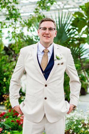 A groom in a crisp white suit with navy vest and tan tie poses in a greenhouse.