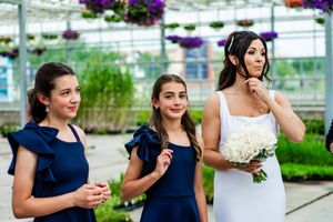 Wedding party members in navy and white dresses pose in a greenhouse with hanging flower baskets.