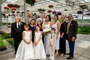 Wedding party members gather in a bright greenhouse filled with hanging plants and flowers.