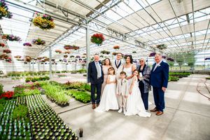 Wedding party gathered in a bright greenhouse filled with rows of plants and hanging flower baskets.
