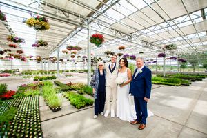 Wedding group photo in a large commercial greenhouse filled with colorful hanging baskets and potted plants.